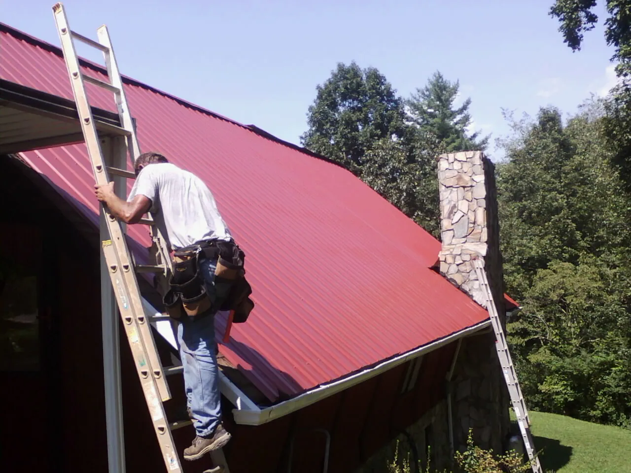 Roofer on a ladder beside a red metal roof and stone chimney — Soffit & Fascia Repair by Roofing Brothers in Monitor